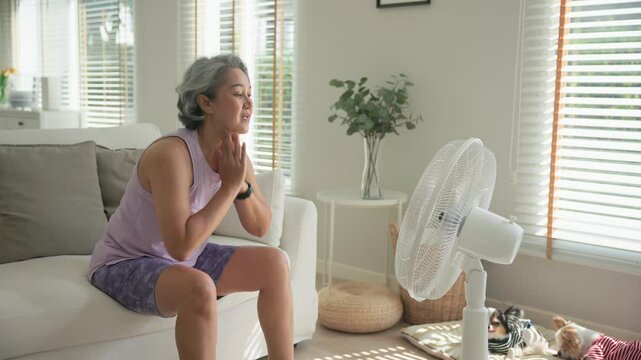 Asia adult people middle age old woman sit at sofa home feel hot flash fever use fan waving hand blowing wind face elder health care summer heat aging issue. Sad pain sweat tired stroke suffer stress.