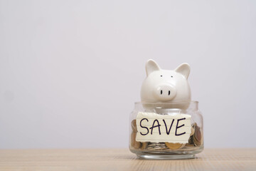 A white ceramic piggy bank sits atop a glass jar filled with coins on a wooden surface, symbolizing...