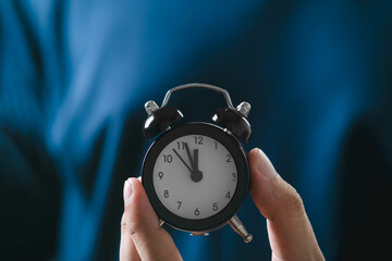 Close-up of hands holding a small black alarm clock in front of a dark blue background, symbolizing...