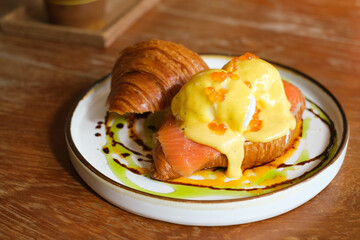 Delicious croissant topped with smoked salmon and creamy poached eggs, garnished with rich hollandaise sauce on a rustic wooden table.