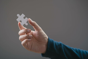 A close-up view of a hand holding a single white puzzle piece against a neutral background, symbolizing completion, curiosity, or problem-solving.