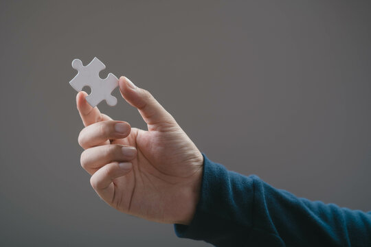 A close-up view of a hand holding a single white puzzle piece against a neutral background, symbolizing completion, curiosity, or problem-solving.