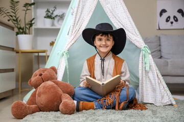 Cute boy dressed as cowboy reading adventure book in play tent at home