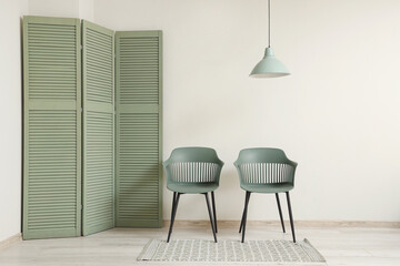 Chairs, folding screen and hanging lamp near white wall in room