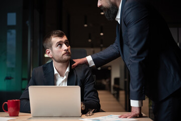 Businessman sitting at a desk with a laptop, looking up at a senior colleague or boss placing a...