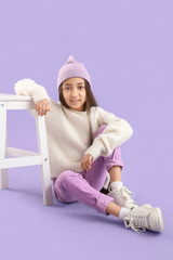Stylish girl in hat sitting near stool on lilac background