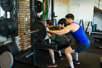 Determined man performing squats with barbell with assistance from his personal trainer during...