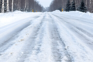 Obraz premium Winter road covered in snow and ice, with tire tracks visible. Trees line the sides of the road in the wintry landscape.