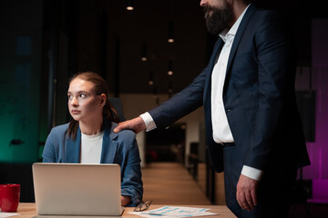 Young woman works on her laptop in a dimly lit office, showing discomfort as a male colleague...