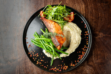 A beautifully plated dish featuring grilled salmon, sautéed green beans, creamy mashed potatoes, and fresh salad with cherry tomatoes, garnished with herbs and spices.
