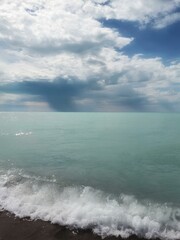 Azure Lake Balkhash with gentle waves and a sandy shore. Above the horizon, a sky-blue sky with large cumulus clouds, with rain falling in places. The vivid colors create a picturesque scene.