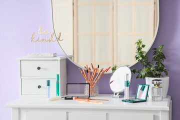 Table with drawers, plant, makeup products and mirror on lilac wall in room