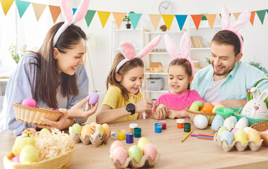 Happy smiling family having fun painting eggs during Easter preparation. Mother, father and two children wearing funny bunny ears sitting at table in kitchen together celebrating Easter holiday.