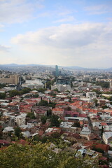 Panoramic view of Tbilisi a city, Georgia blending historic architecture with modern skyscrapers. Red-roofed buildings spread across the landscape, with green hills and a blue sky in the background