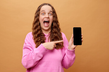 A joyful woman with wavy hair and blue eyes enthusiastically points at a smartphone she holds in...