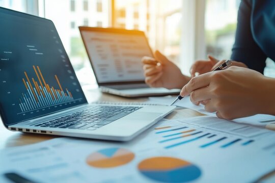Data charts on laptops. Hands pointing to graphs. Analyzing business statistics. Finance and data analysis. Teamwork reviewing charts. Documents and technology.