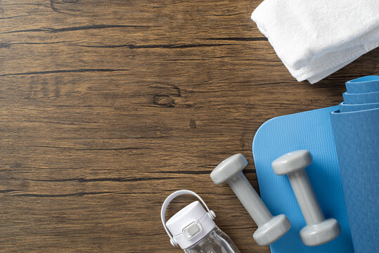 Workout equipment including a yoga mat, dumbbells, water bottle, and towel on wooden surface