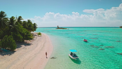 Lone woman walks along a pristine pink sand beach on a remote tropical island, with small boats...