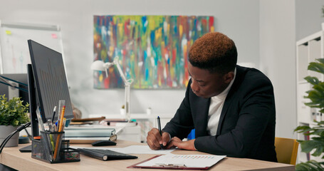 Dark-skinned man in black suit sits at desk in front of computer monitor, describes graphs printed...