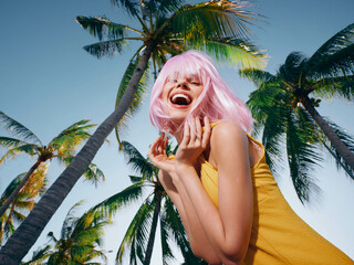 Pinkhaired woman in yellow dress stands confidently with hands on hips, smiling in front of lush palm trees