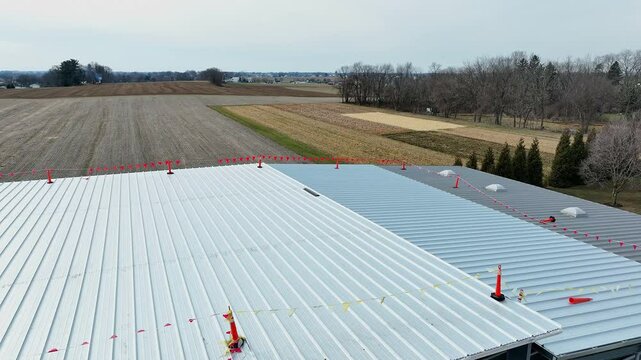 Workers are installing a new metal roof on an agricultural building. The landscape features expansive fields and trees, showcasing rural life. The process emphasizes safety with cones and markings.