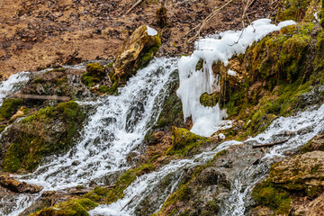 A beautiful multi-tiered waterfall flows over moss-covered rocks in a forest setting. The long...