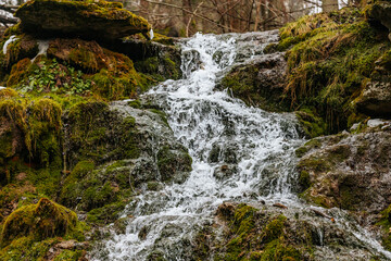A beautiful multi-tiered waterfall flows over moss-covered rocks in a forest setting. The long exposure captures the smooth movement of the water, creating a peaceful and serene natural scene.