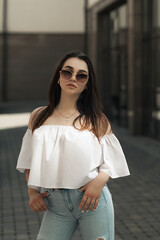 a woman stands on the street on a sunny day. She is wearing a white off-the-shoulder blouse and blue jeans. She's wearing sunglasses and looking straight into the camera
