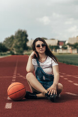 A girl sits on a stadium treadmill. She is wearing a white T-shirt with stripes and denim overalls....