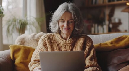 Happy relaxed 55 years old woman sitting on sofa at home looking at laptop. Smiling middle aged lady working in living room at home. Mature older female doing job on couch enjoying chill. High quality