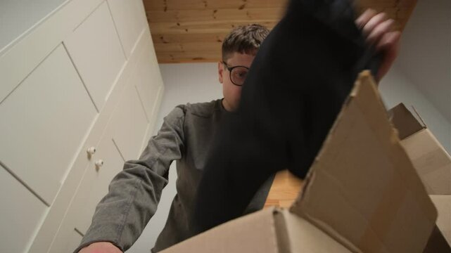 A young man wearing glasses anxiously unpacks clothes from a cardboard box, pulling out items with frustration. The disorganized space filled with boxes suggests an unplanned move or forced relocation