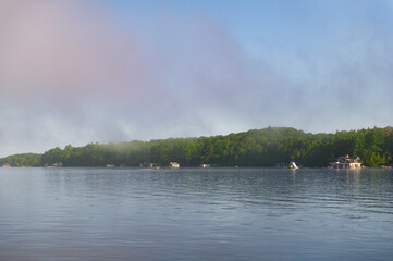 Lake Joseph in Muskoka, Ontario, Canada. An early morning scene with low clouds hovering above the calm water, illuminated by soft orange tones. Cottages are visible along the distant shore.
