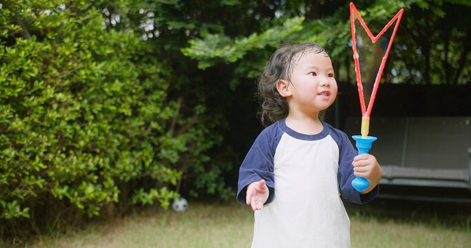 A little Asian child enjoys Playing blowing Colorful soap bubbles Having Fun in a Lush Green Garden surrounded by greenery and sunlight on a Sunny Day, capturing the essence of carefree childhood fun.