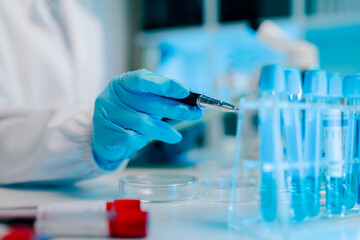 Scientist is holding a small device in a lab with a blue liquid in a beaker. The lab is well lit and the scientist is wearing a white lab coat