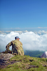 A man in military clothes sits on a stone mountain above the clouds