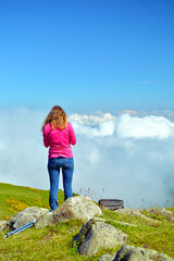 Young woman with pink jacket standing on mountain stone and looking at clouds