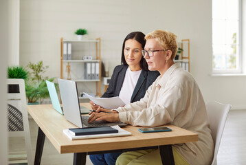 Two female colleagues sitting at desk in office, using laptops and working with documents. Company employees discussing business strategy or financial results, analyzing accounting data together.