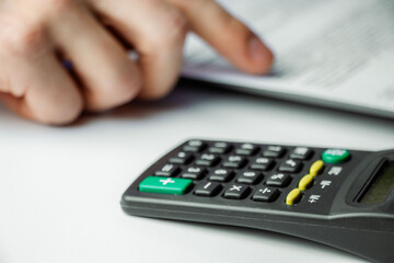 A person calculates finances using a black calculator on a white desk. A printed document with text...