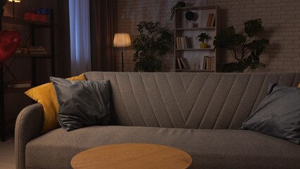 Close shot of living room in cozy apartment, modern sofa standing in the middle of the room with colorful pillows, low light and modern furniture creating stylish atmosphere.