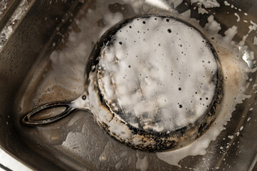 A heavily burnt cast iron pan being cleaned in a stainless steel sink, covered in soap and grime....