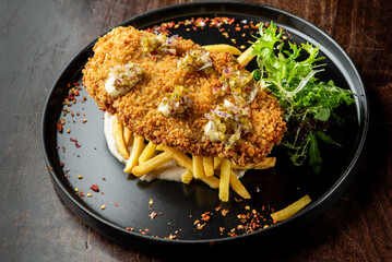 A beautifully plated crispy fried fish on a bed of golden French fries, garnished with fresh greens and drizzled with a tangy sauce, presented on a dark plate.