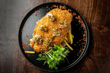 A beautifully plated crispy fried fish on a bed of golden French fries, garnished with fresh greens and drizzled with a tangy sauce, presented on a dark plate.