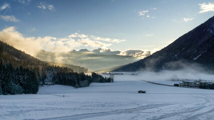 Mountain valley shrouded in mist at sunset in winter. Thick mist envelops the valley at dusk, mountains picks on horizon,  clouds on blue sky. South Tyrol, Trentino Alto Adige, Alps, Italy, Europe.