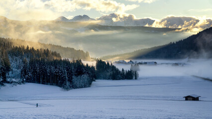 Mountain valley shrouded in mist at sunset in winter. Thick mist envelops the valley at dusk, mountains picks on horizon,  clouds on blue sky. South Tyrol, Trentino Alto Adige, Alps, Italy, Europe.