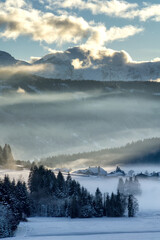 Mountain valley shrouded in mist at sunset in winter. Thick mist envelops the valley at dusk, mountains picks on horizon,  clouds on blue sky. South Tyrol, Trentino Alto Adige, Alps, Italy, Europe.
