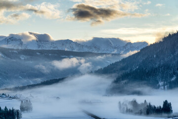 Mountain valley shrouded in mist at sunset in winter. Thick mist envelops the valley at dusk, mountains picks on horizon,  clouds on blue sky. South Tyrol, Trentino Alto Adige, Alps, Italy, Europe.