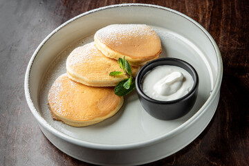 Three fluffy pancakes dusted with powdered sugar are served on a white plate, accompanied by a small black bowl of creamy yogurt and a fresh mint leaf.