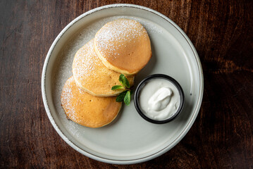Three fluffy pancakes dusted with powdered sugar are served on a white plate, accompanied by a small black bowl of creamy yogurt and a fresh mint leaf.