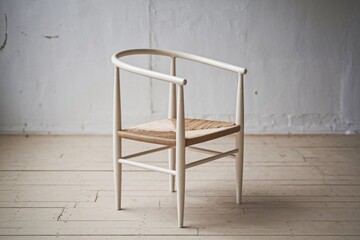A minimalist white wooden chair with a woven seat, positioned against a textured, neutral wall and wooden floor.