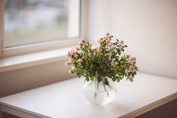 Small pink roses harmonize with lush green leaves in a glass vase, creating an inviting atmosphere on a minimalist white desk beside a bright window during the day